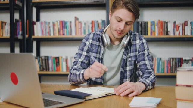 Young man studying in library with laptop, headphones, and notebook during study session for academic success