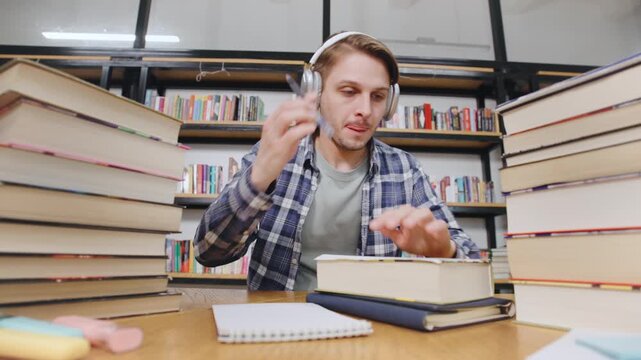 Student studying in a library with stacks of books and headphones on while taking notes at a wooden table during the day