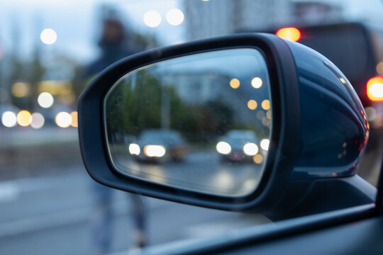 Blurry nighttime street reflection in a car side mirror showing approaching traffic headlights and a pedestrian, emphasizing urban travel and safety
