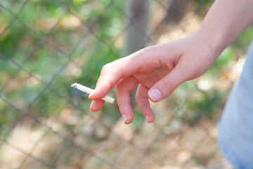 Female hand holding cigarette in front of the fence. Focus on cigarette.