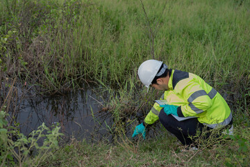 Environmental engineer wearing safety gear collecting water sample from polluted pond for contamination analysis, environmental testing, and ecological impact assessment in natural outdoor setting.