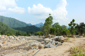 A wide landscape view of a dry riverbed or stream with scattered gray boulders and sandy banks, backed by lush green mountain slopes and a bright blue sky with white cumulus clouds © Fazal