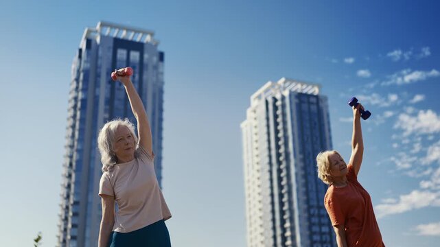 Two senior ladies exercising with dumbbells against city skyscrapers in park