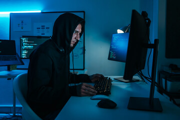 Caucasian male hacker in black hoodie working on computer in dark office, attempting data breach and cybersecurity hacking.