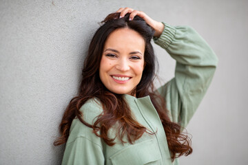 Naklejka premium Happy woman smiling at camera with hand in hair leaning on wall