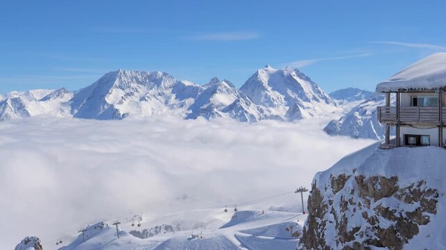 Alpine panorama with snow capped peaks rising above sea of clouds. Chairlift operates at popular ski resort on sunny day. Showcasing breathtaking winter wonderland. Mountain chalet on edge of cliff