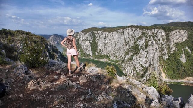 Woman standing on a rocky viewpoint, admiring the scenic Danube river winding through the rugged gorge and towering cliffs on the border of Serbia and Romania, enjoying nature and a clear sky