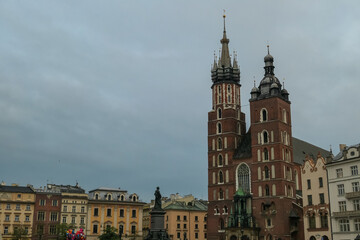 The majestic St. Mary's Basilica dominates the skyline of Krakow's historic Main Market Square, surrounded by traditional tenement houses and a famous monument under a cloudy, overcast sky.