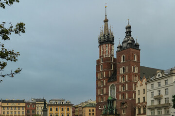 Obraz premium St. Mary's Basilica stands as a historic landmark in Krakow's Main Market Square, surrounded by colorful old town buildings and the Adam Mickiewicz monument under a cloudy sky in Poland.