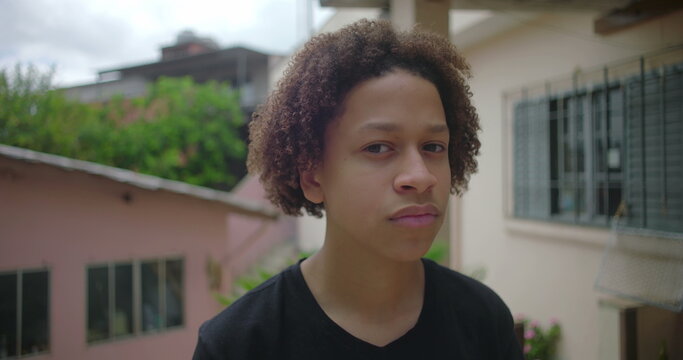 Latino teenage boy from African descent with a serious expression standing outdoors in a Latin American residential neighborhood, conveying thoughtfulness and introspection - Powered by Adobe