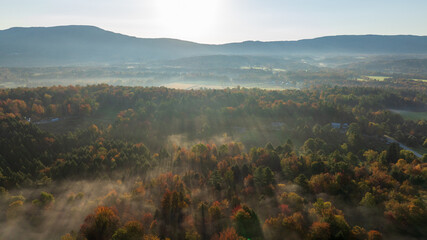 Sunbeams break through fog over a colorful autumn forest in a misty Vermont valley at sunrise.