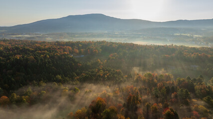 Sunbeams break through fog over a colorful autumn forest in a misty Vermont valley at sunrise.