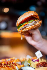 Close-up of a hand holding a juicy hamburger with beef patty, lettuce, tomato, pickles, and dripping sauce. Loaded fries and tortilla chips in background. Restaurant setting.