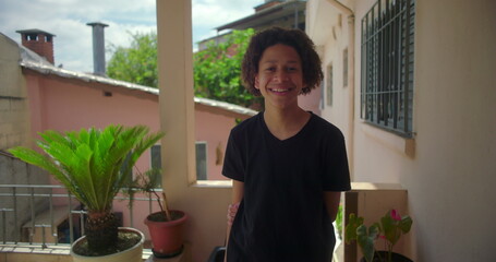 Mixed-race curly-haired teenager standing confidently on a porch in a warm, tropical neighborhood surrounded by greenery, cheerful expression and relaxed posture