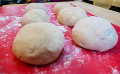 Six round balls of fresh, raw dough are resting on a red surface dusted with white flour. The smooth portions are proofing, getting ready for the next step in the baking process for bread or pizza.