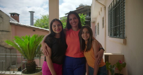 Smiling mother standing with her two daughters on a porch in a South American neighborhood,...
