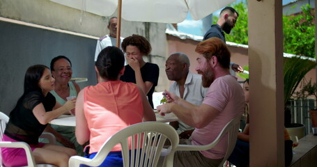 Diverse group of people enjoying outdoor barbecue gathering, sharing food and laughing together under patio umbrella in backyard © Marco