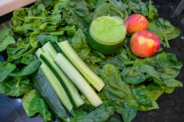 A nutritious green smoothie is presented in a glass, surrounded by its fresh ingredients of leafy spinach, sliced cucumber, and crisp apples, embodying a healthy lifestyle in a home or restaurant.