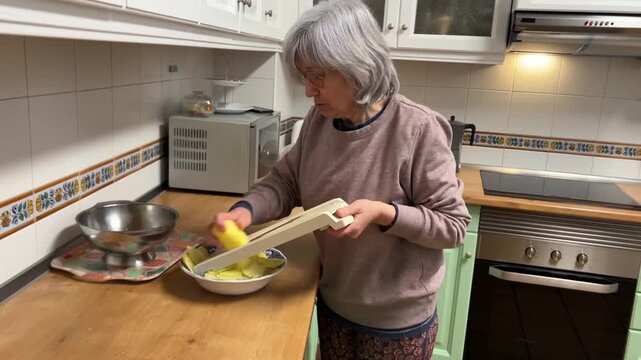 Senior woman with white hair slicing potatoes with mandoline slicer in kitchen preparing Spanish omelette