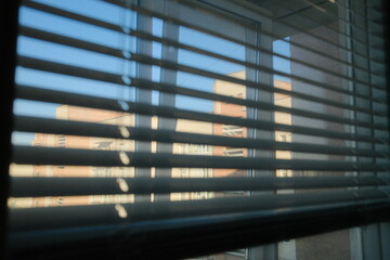 View through blinds shows exterior of building and blue sky; sunny day.