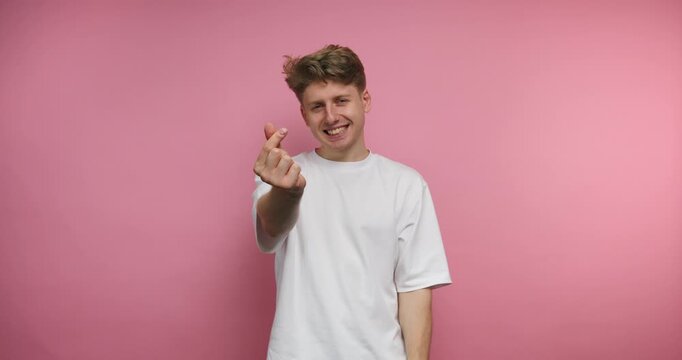 footage of young playful man forms a heart gesture with his fingers, showcasing warmth and affection on pink background