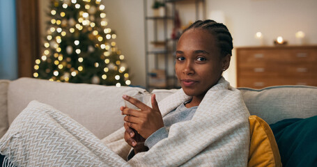A person of multi-racial background sits cozily on a couch, wrapped in a blanket. They take a sip...