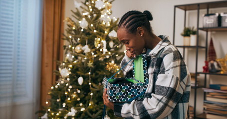A young student of multi-ethnic background stands by a Christmas tree, unwrapping a gift. She unties the blue ribbon, opens the box, and finds a warm black hat. Smiling, she hugs it happily.