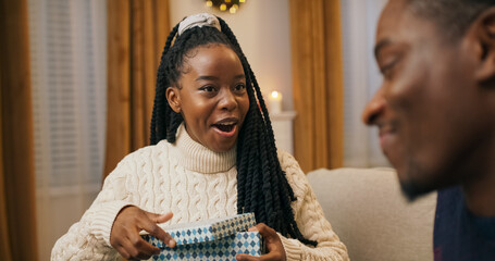 The close-up shows a young man handing a gift to his friend. The young woman of multi-ethnic background unties the ribbon, opens the box, and is pleasantly surprised. Both friends are smiling and