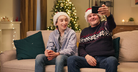 Grandson and grandfather are sitting on couch in the living room. They have video call on phone with relatives, talking, smiling. They are wearing Santa's hats. A Christmas tree flickers in the room.