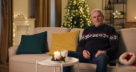 Fototapeta na wymiar Sitting on a sofa in the living room on Christmas Eve, a lonely middle-aged man dropped his Santa's hat in disappointment. Left alone today, he set the hat aside and wears a warm sweater.