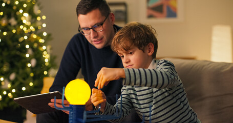 A son and dad play with a model of the planetary system on a couch. The man is explaining the construction of the cosmos to the boy, using the toy as an example and naming the planets.