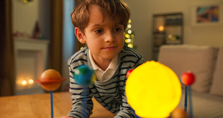Fototapeta premium A close-up shows a 7-year-old boy looking at a child's model of the solar system. He wears a shirt and a striped sweater, looking at the planets with interest and a smile. A Christmas tree flickers