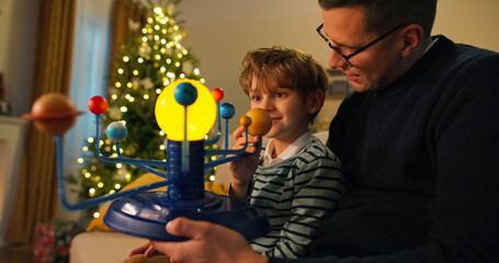 A dad with glasses and his son sit on a couch. The dad holds a model of the solar system, showing it to his son, who looks with interest and smiles. A shimmering Christmas tree is behind them.