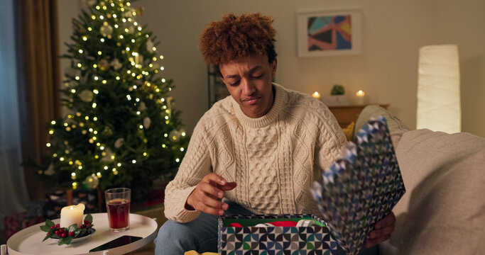 A curious man, who is also smiling and handsome, sits on a couch near a Christmas tree. In a white sweater, he opens a gift box, only to feel disappointed and sad because he doesn't like the surprise.