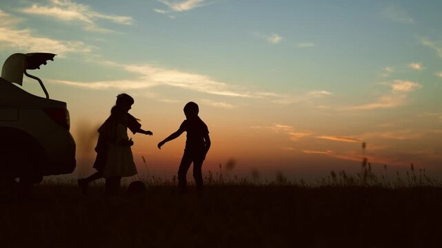Kids playing at sunset by car, silhouettes. Children play football with ball have fun in nature field park. Cheerful joyful kids girl boy siblings playing games together. Active lifestyle leisure time