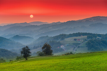 Glowing sunset illuminating misty rolling hills with scattered rural houses, green meadows, and...