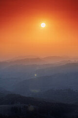 Fototapeta premium Sun setting orange sky above layered rolling hills with atmospheric haze. Capturing serenity and natural beauty at dusk