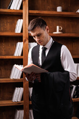 Elegant young man in black suit posing in a modern studio interior with bookshelves and leather sofa, stylish business portrait