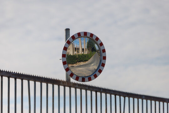 Convex mirror reflects a road leading to damaged buildings and lush green bushes under a cloudy sky.