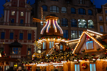 Illuminated Christmas carousel and wooden pyramid with warm golden bokeh lights at night holiday market in historic European town square. Blurred festive entertainment background shot.