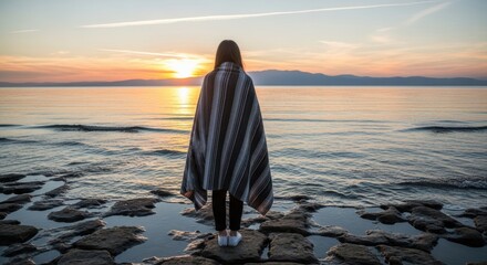 Female wrapped in blanket watching sunset over serene lake with rocky shoreline