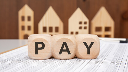 Wooden cubes spelling PAY on financial spreadsheet with wooden house models in blurred background