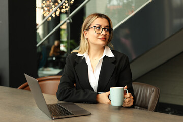 A girl in an office is drinking coffee while working on a laptop.