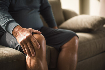 Man seated with hand on knee suggesting pain or discomfort. Seeking relief or assessing injury indoors on the couch.