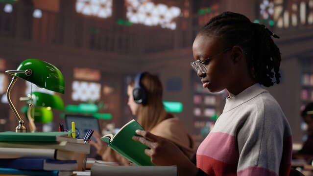 African american girl reading and taking notes for exam at the college, studying political science for upcoming class lesson in a library at university. Young student consults textbooks. Camera A. - Powered by Adobe