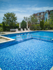 Clean outdoor pool with clear blue water, sun loungers, and umbrellas near modern apartment building. Peaceful summer vibe and relaxation area.