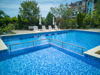 Clean outdoor pool with clear blue water, sun loungers, and umbrellas near modern apartment building. Peaceful summer vibe and relaxation area.