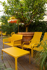 A cozy lounge area in a cafe on an open veranda with yellow chairs and a table. Lush green plants surround the space, creating a relaxing atmosphere.