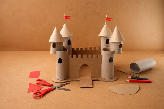 Handmade cardboard castle with red paper flags and crafting tools on table. DIY art project made of paper rolls and recycled materials. Studio composition for design and print. Top view,