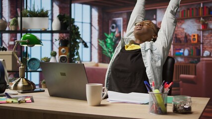 Woman in home office stretches back, feeling fatigued while working on laptop. Remote employee in apartment feeling backpain, relieving pain, solving tasks on notebook, camera A